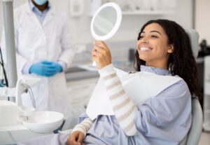 A woman with dark curly hair smiles while looking into a hand mirror in a dental clinic, feeling as satisfied as seeing positive results on an SEO pipeline. The dentist nearby wears gloves, a white coat, and a face mask.