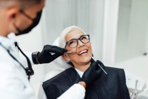 A smiling older woman with short gray hair and glasses sits in a dental chair while a dentist in gloves and a mask examines her teeth, much like an expert reviewing data on an SEO pipeline.